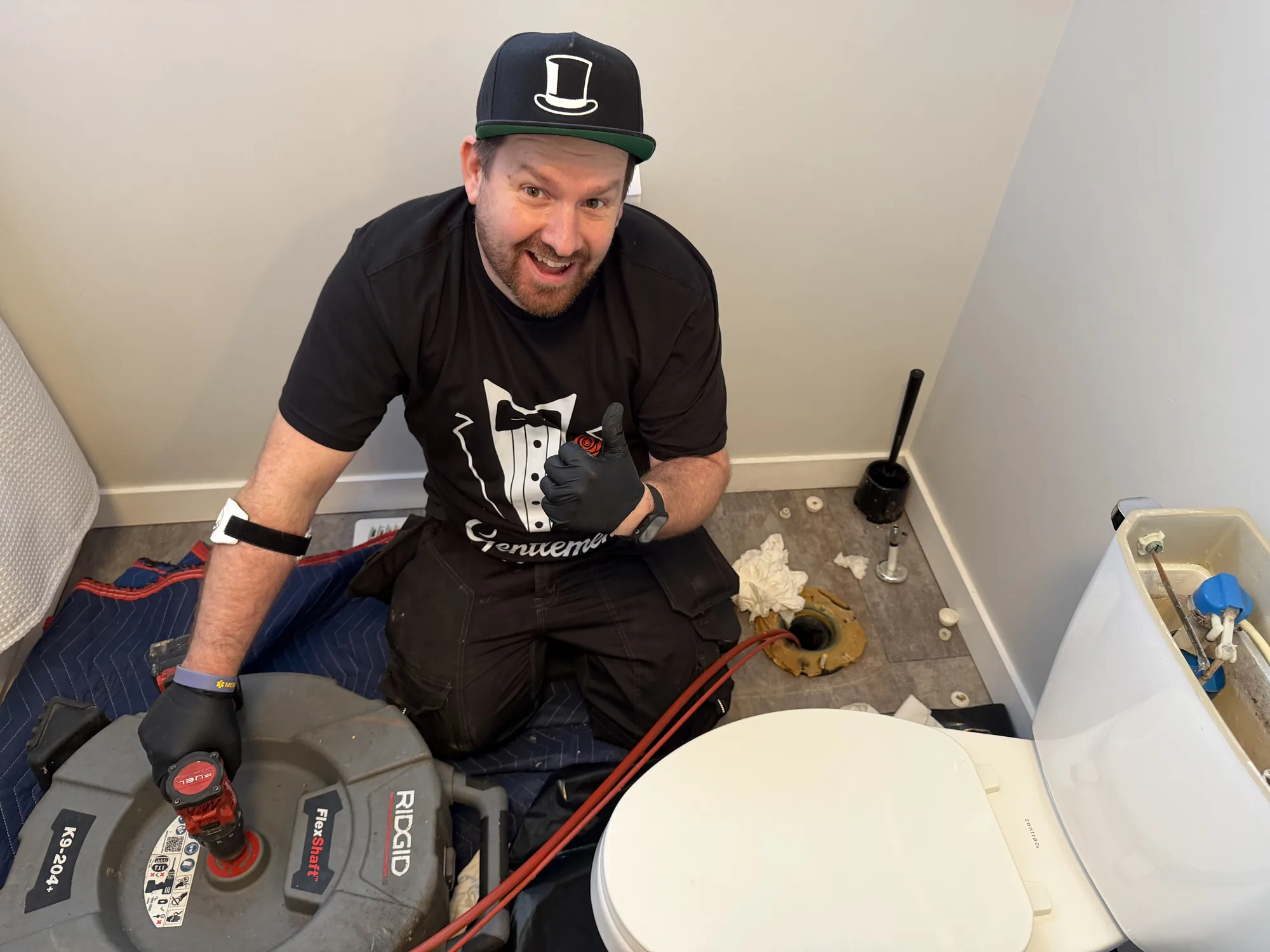 Plumber kneeling beside an exposed toilet flange with a drain machine.
