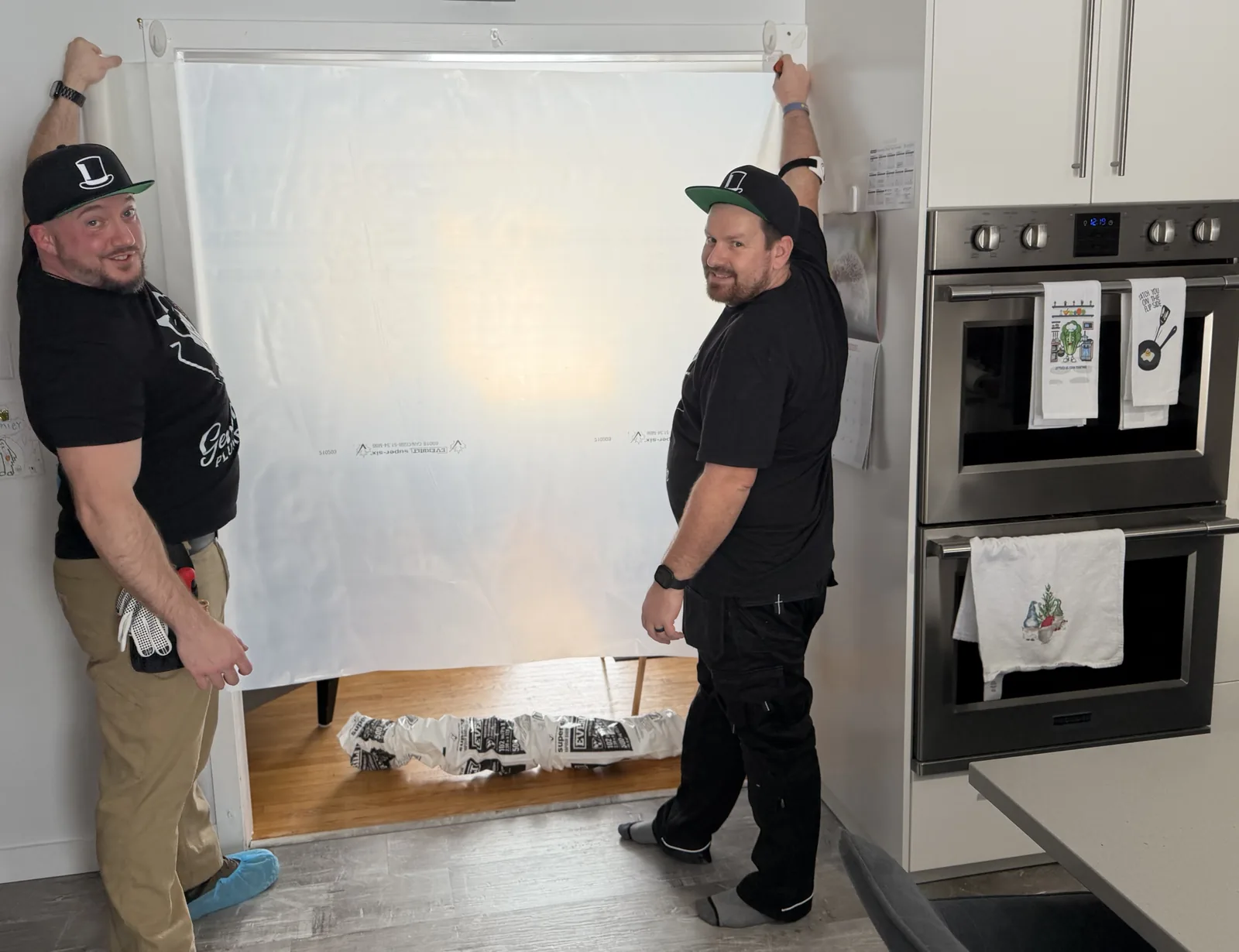 Two Gentlemen Plumbing technicians holding a clear dust barrier across a kitchen doorway.